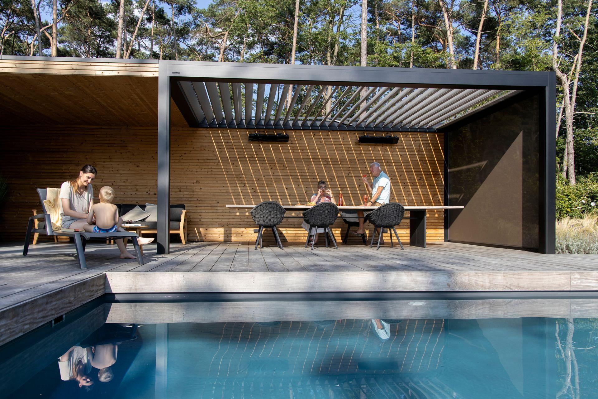 People relaxing in a modern wooden outdoor pavilion with a pool and forest backdrop.