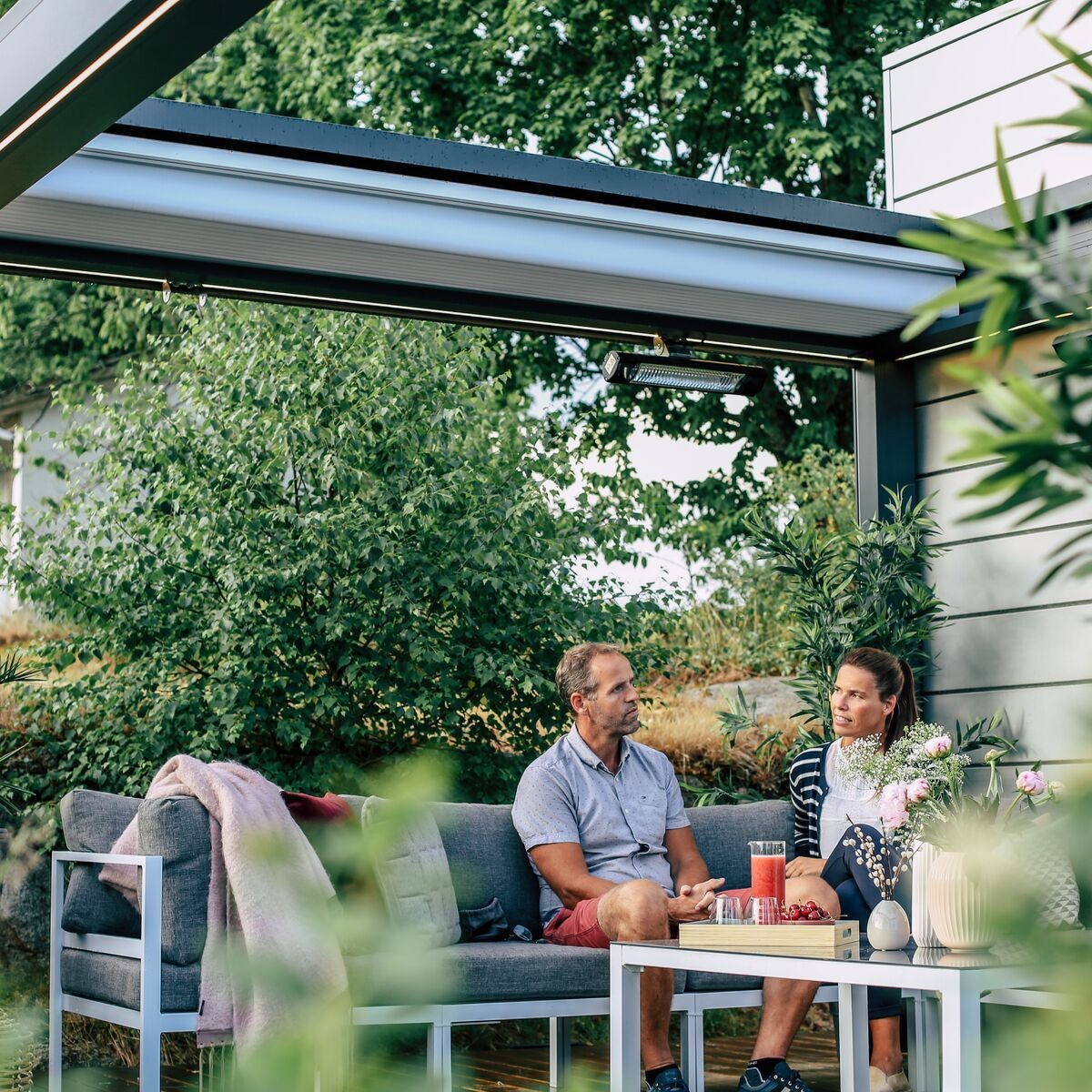 Couple sitting on outdoor sofa under pergola, surrounded by greenery and flowers.