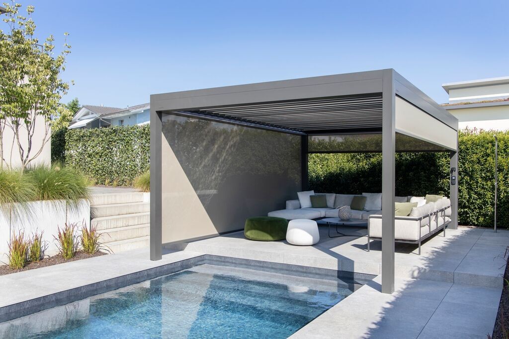 Modern outdoor pergola with lounge seating beside a swimming pool, surrounded by hedges and greenery.