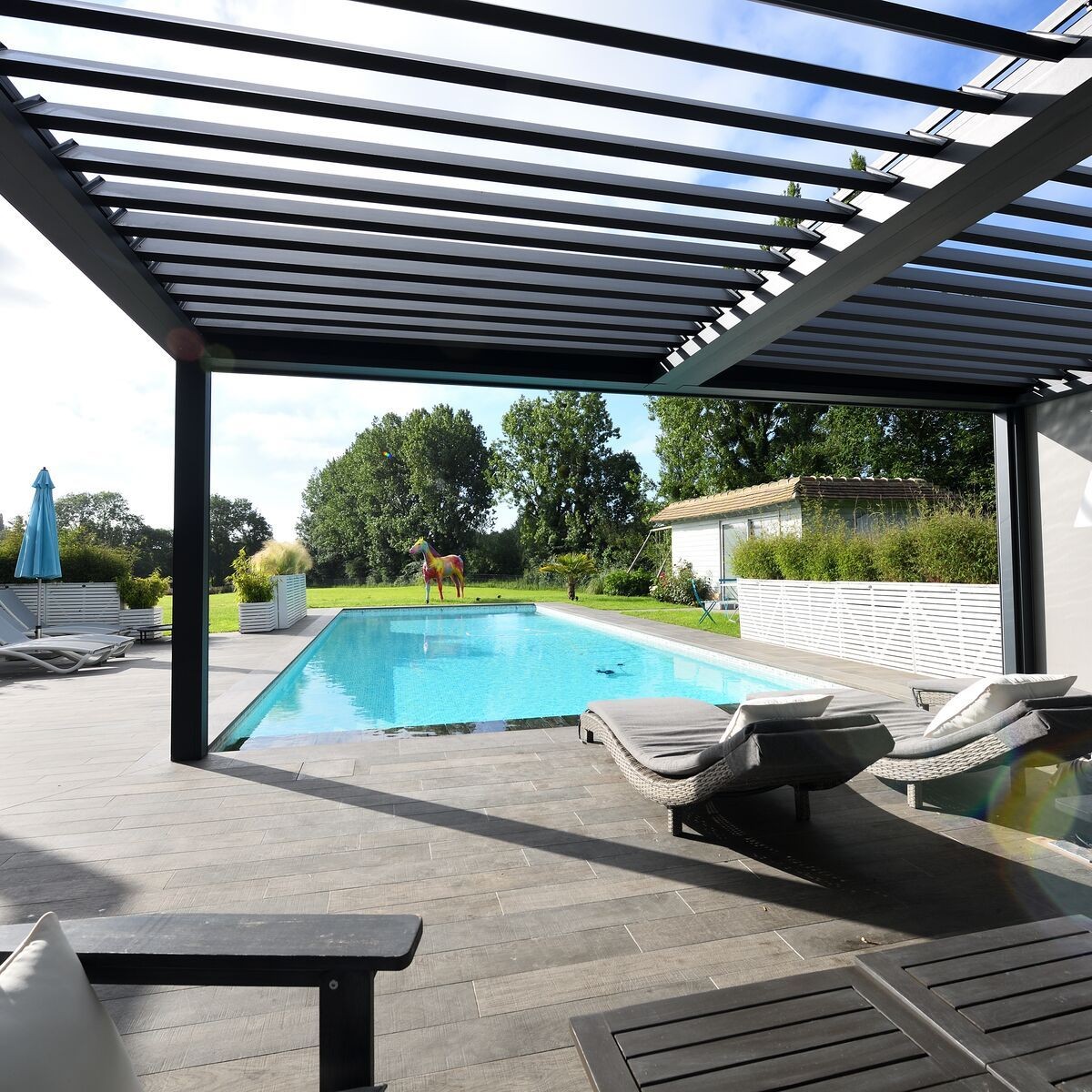 Modern patio with loungers overlooking a pool and garden sculpture in sunny weather.