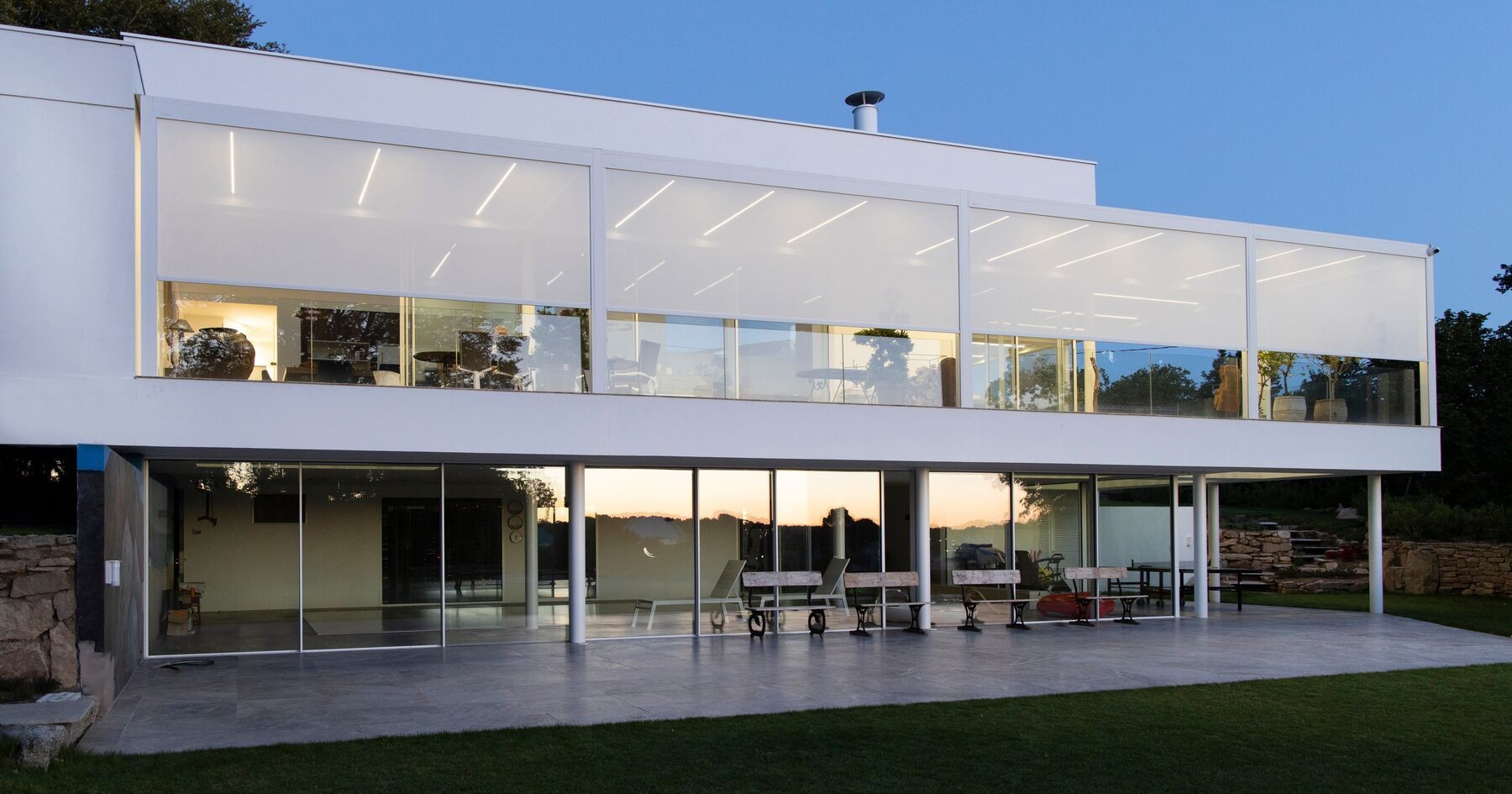 Modern two-story house with large glass windows and a lush green lawn at dusk.