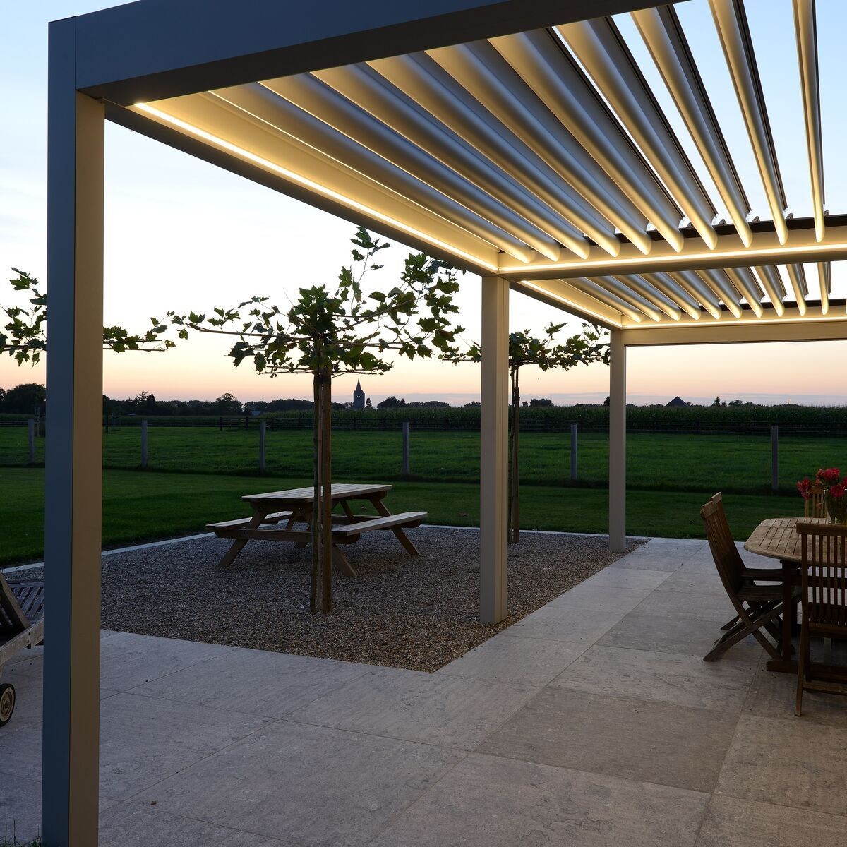 Outdoor patio with modern pergola and wooden tables overlooking a green field at sunset.