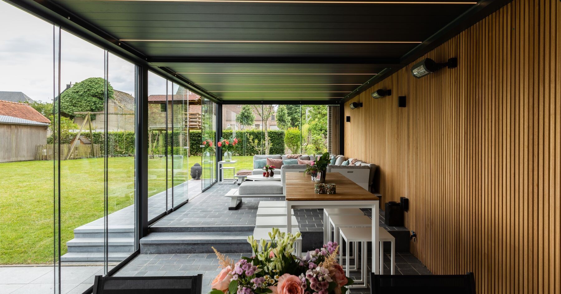 Modern outdoor patio with glass walls, wooden panels, and seating, overlooking a garden.