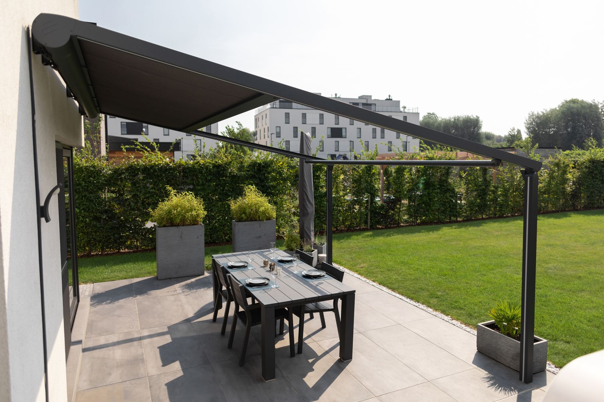 Outdoor patio with dining table under a modern awning, surrounded by greenery.