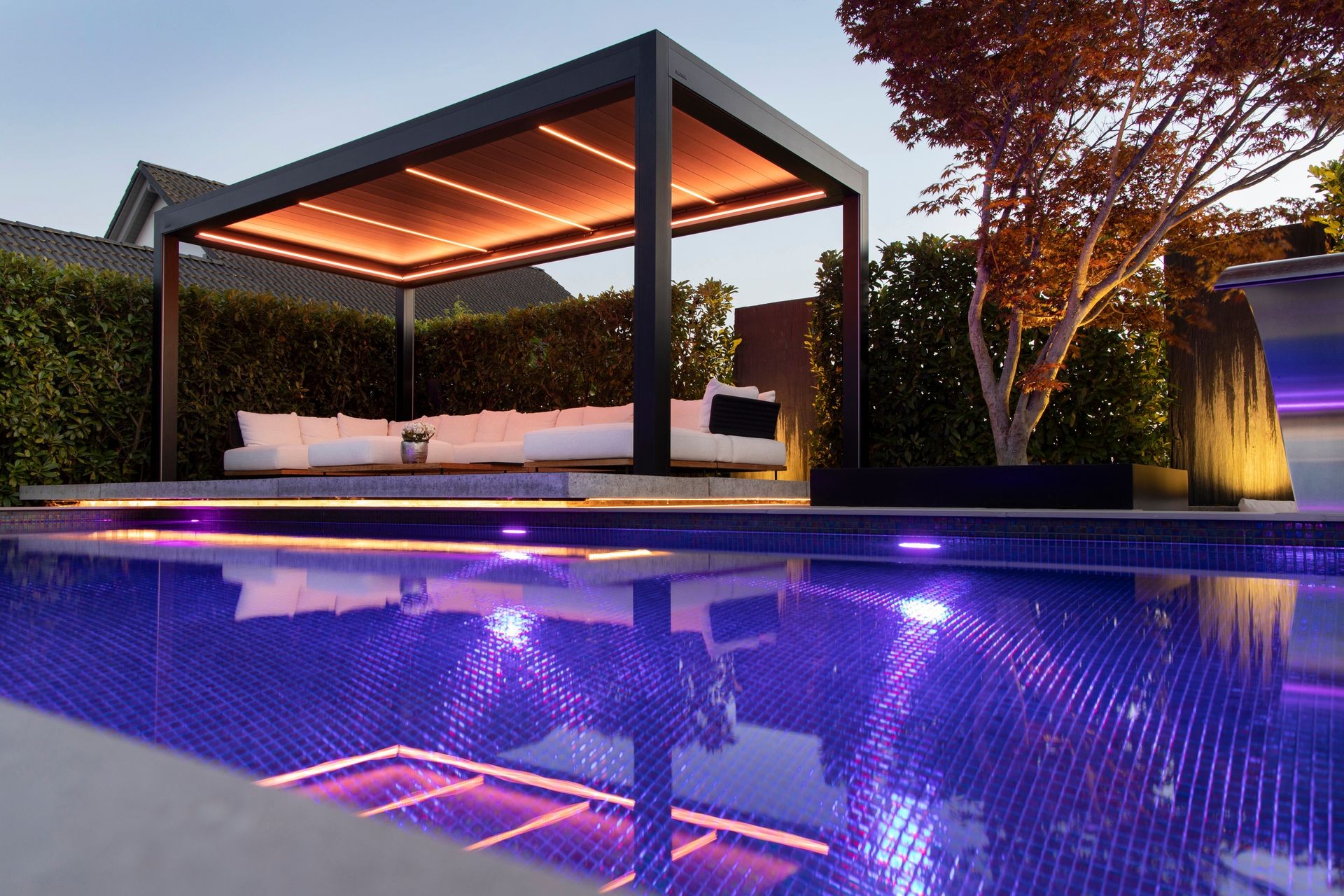 Luxury outdoor seating area with modern cabana and pool illuminated by ambient lights at dusk.