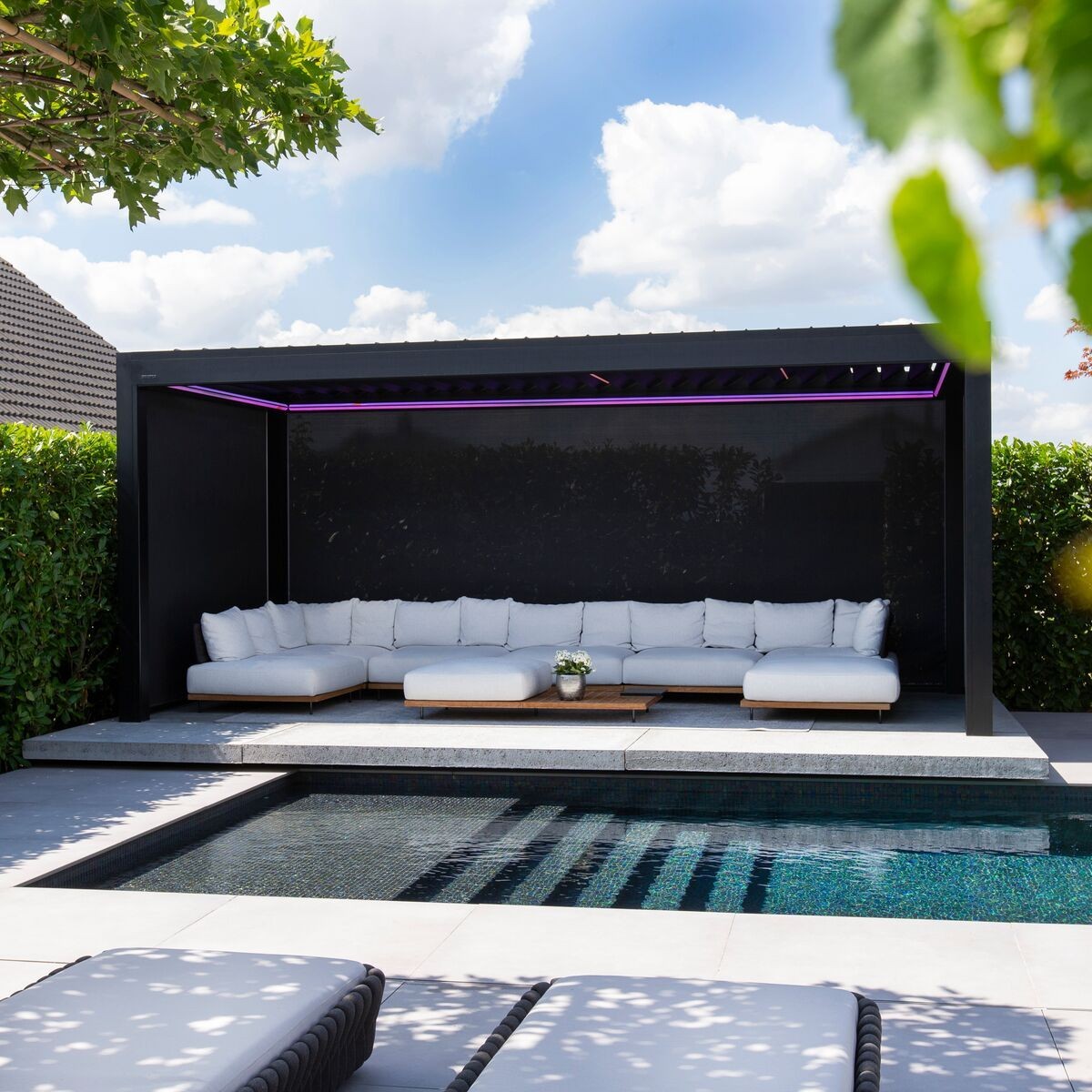 Outdoor lounge area with white sofas by a pool, under a black canopy, surrounded by greenery.