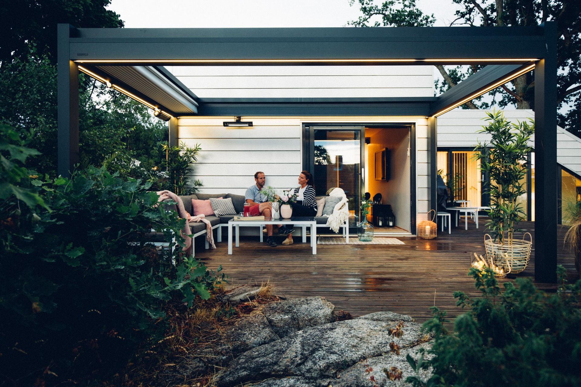 Couple sitting on a patio with modern furniture and outdoor lighting, surrounded by greenery.